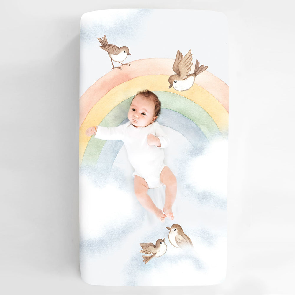 Baby crib sheet with watercolor rainbow design, light blue background, birds and clouds