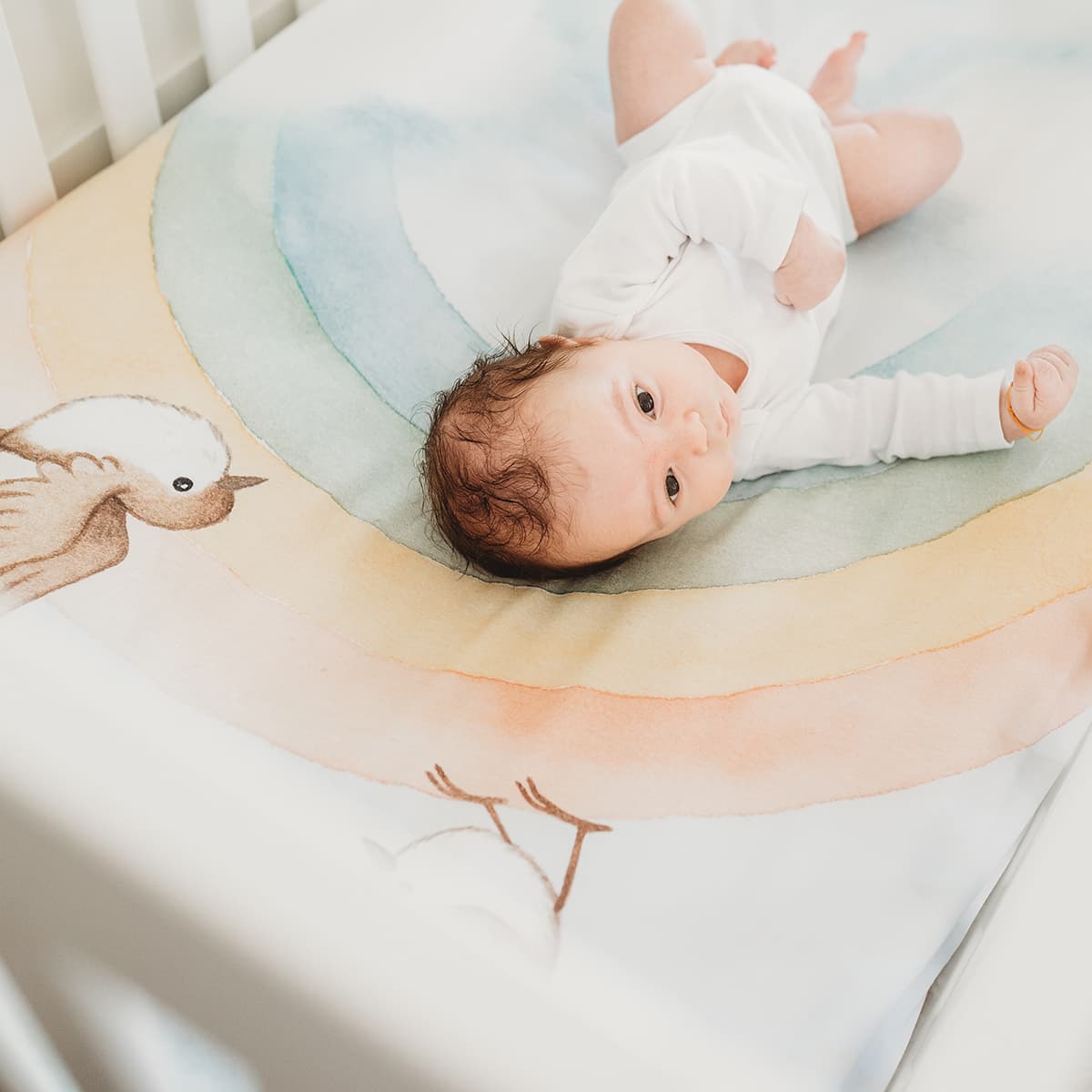 Baby crib sheet with watercolor rainbow design, light blue background, birds and clouds