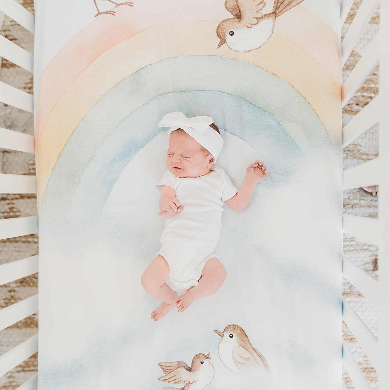 Baby crib sheet with watercolor rainbow design, light blue background, birds and clouds